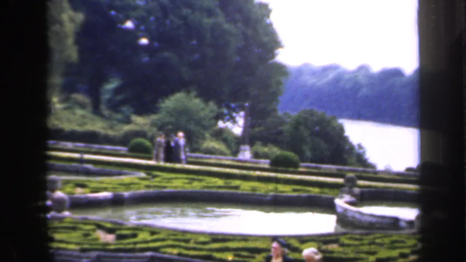 1949 England garden view with formal hedges, a stone fountain basin, visitors on the terrace, and a broad river beyond the trees.