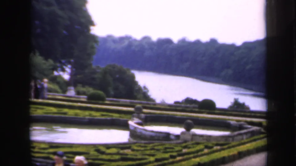1949 England home-movie frame looking across clipped garden beds and a stone basin toward a river and wooded hillside.