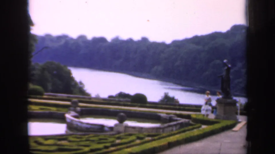 1949 England frame with clipped hedges, a fountain basin, river scenery, a statue on a pedestal, and two visitors at the path edge.