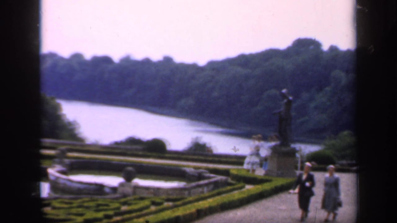 1949 England home-movie view of estate gardens with clipped hedges, a river or lake beyond, a statue, and visitors walking nearby.