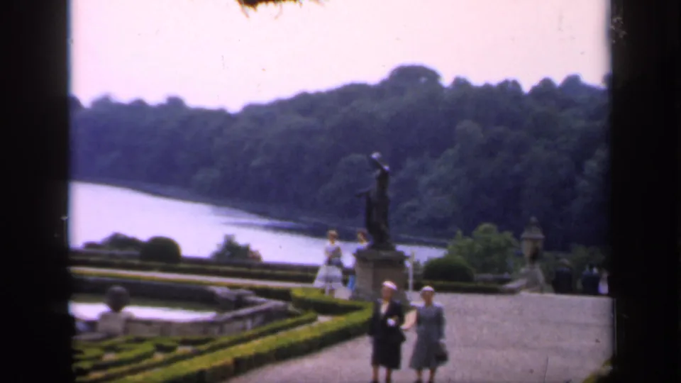 Estate garden scene in England with a statue, clipped hedges, stone terraces, distant water, and visitors in period dress.