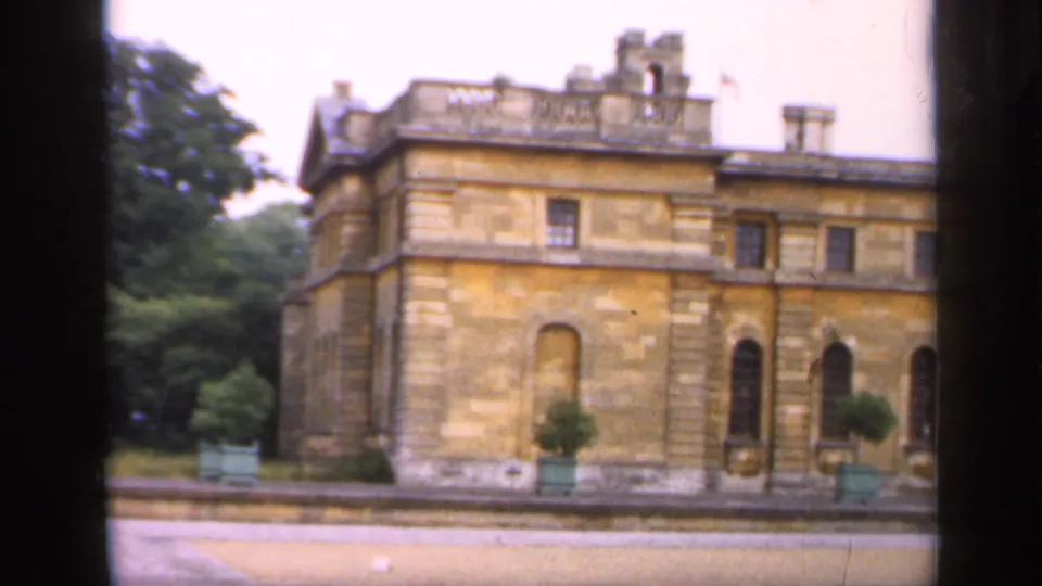 1949 England footage shows a stately stone building facade with arched windows, roofline chimneys, planters, trees, and benches.