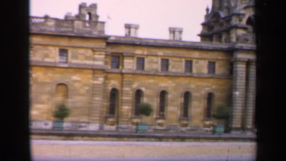 A stately English stone facade fills the frame, showing repeated arched windows, columns, planters, and a pale paved area in front.