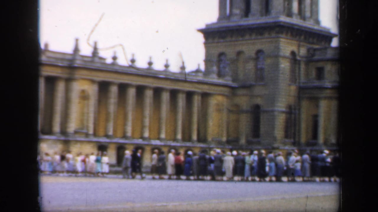 1949 England home-movie frame showing visitors waiting in a long line before a grand classical palace facade.