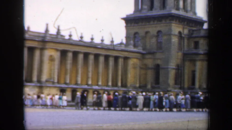 1949 England home-movie frame showing visitors waiting in a long line before a grand classical palace facade.