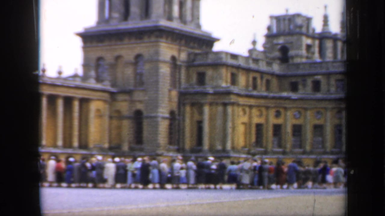 A long public queue stands along the forecourt of a historic palace in England, filmed in soft 8mm color.