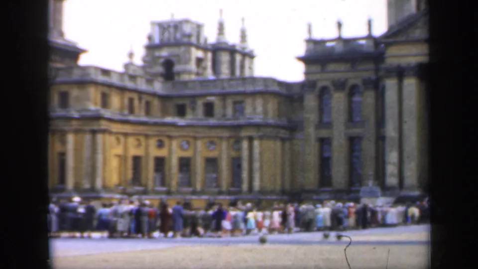 Visitors in period dress wait outside a grand English palace, with towers, columns, and decorative roofline visible.