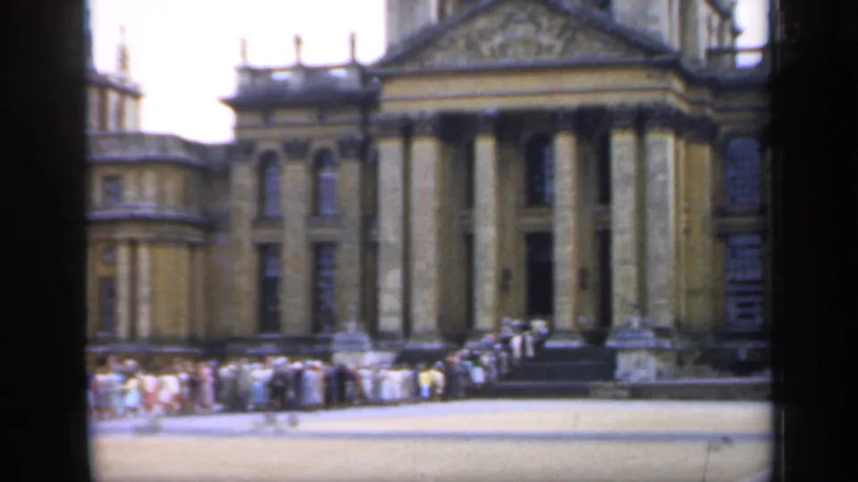 The shot reaches a columned entrance and steps while visitors remain gathered across the palace forecourt.