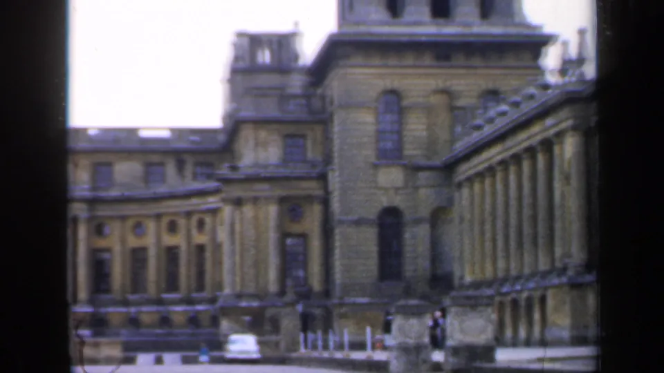1949 England home-movie frame showing a corner view of the monumental stone building, with rows of columns, arched windows, and a small car in front.