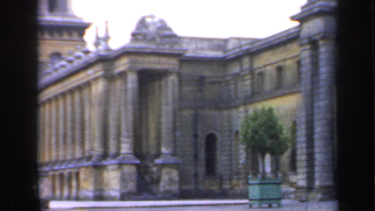 Classical stone building facade with columns, arched doorway, and a green planter partly obscured by dark bars.