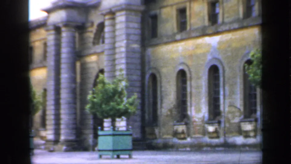 1949 England 8mm frame of a weathered institutional building facade with arched windows, stone columns, and a tree in a green planter.