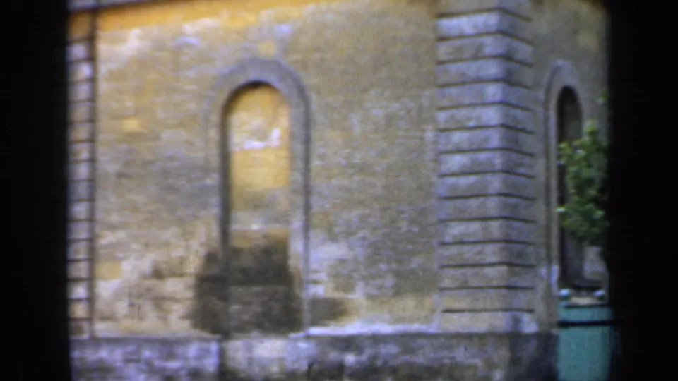 1949 England 8mm frame of a stone institutional facade with an arched opening, column masonry, and part of a green planter at frame edge.
