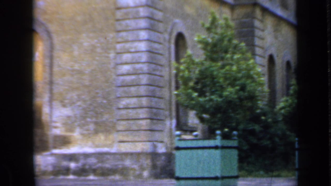 Stone building exterior with arched windows and green planters beside a paved area.