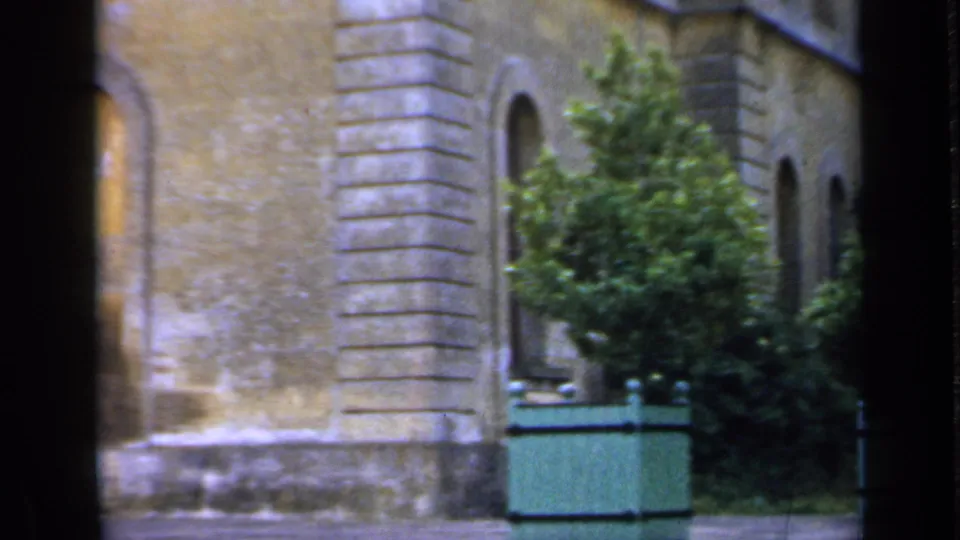 1949 England 8mm frame showing a stone building facade with arched windows, a shrub, and green planters in soft color.