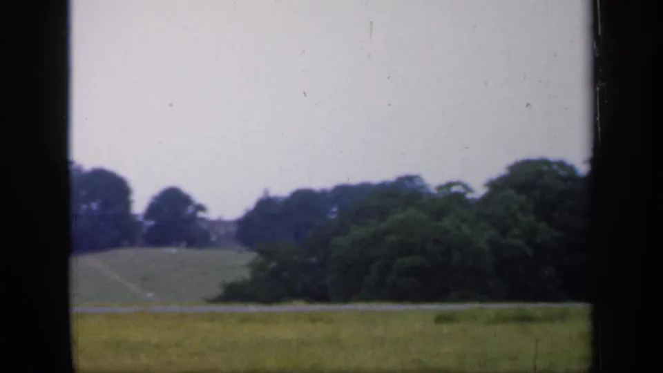 1949 England 8mm view of open grassland, distant tree-lined hills, and a dense cluster of trees at the right edge.
