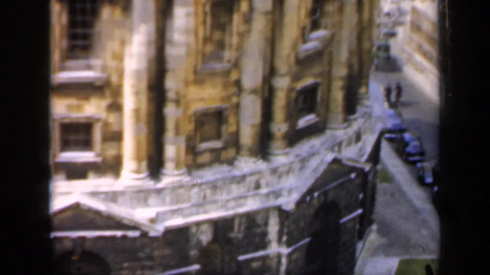 1949 England 8mm frame showing the side of a historic stone building above a narrow street with parked cars.