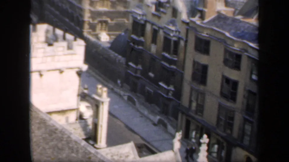Elevated home-movie frame of English historic building facades, a stone corner detail and pedestrians or cars far below.