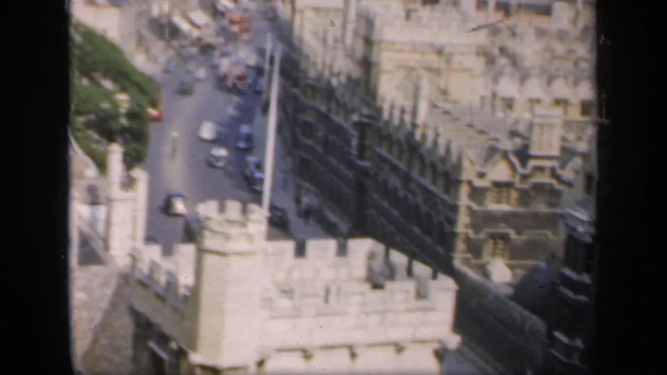 1949 England frame showing a narrow street with cars, pale stone architecture and a long dark roofline from above.