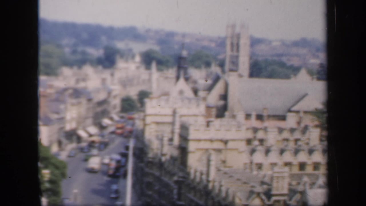 Elevated 1949 England town view with stone facades, a church-like tower, rooftops, and traffic below.
