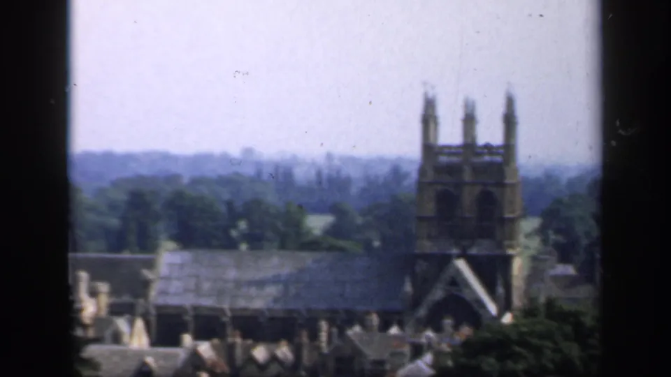 1949 England 8mm view of a Gothic church tower above rooftops, trees, and distant haze, partly divided by a window frame.