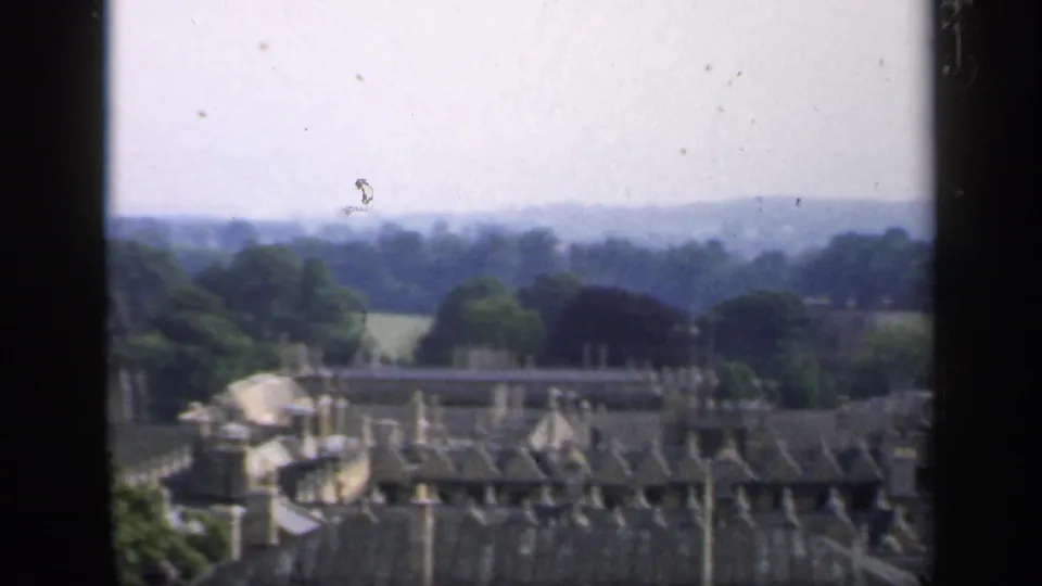 The pan moves across English rooftops and chimney pots toward trees, open fields, and a soft distant horizon.