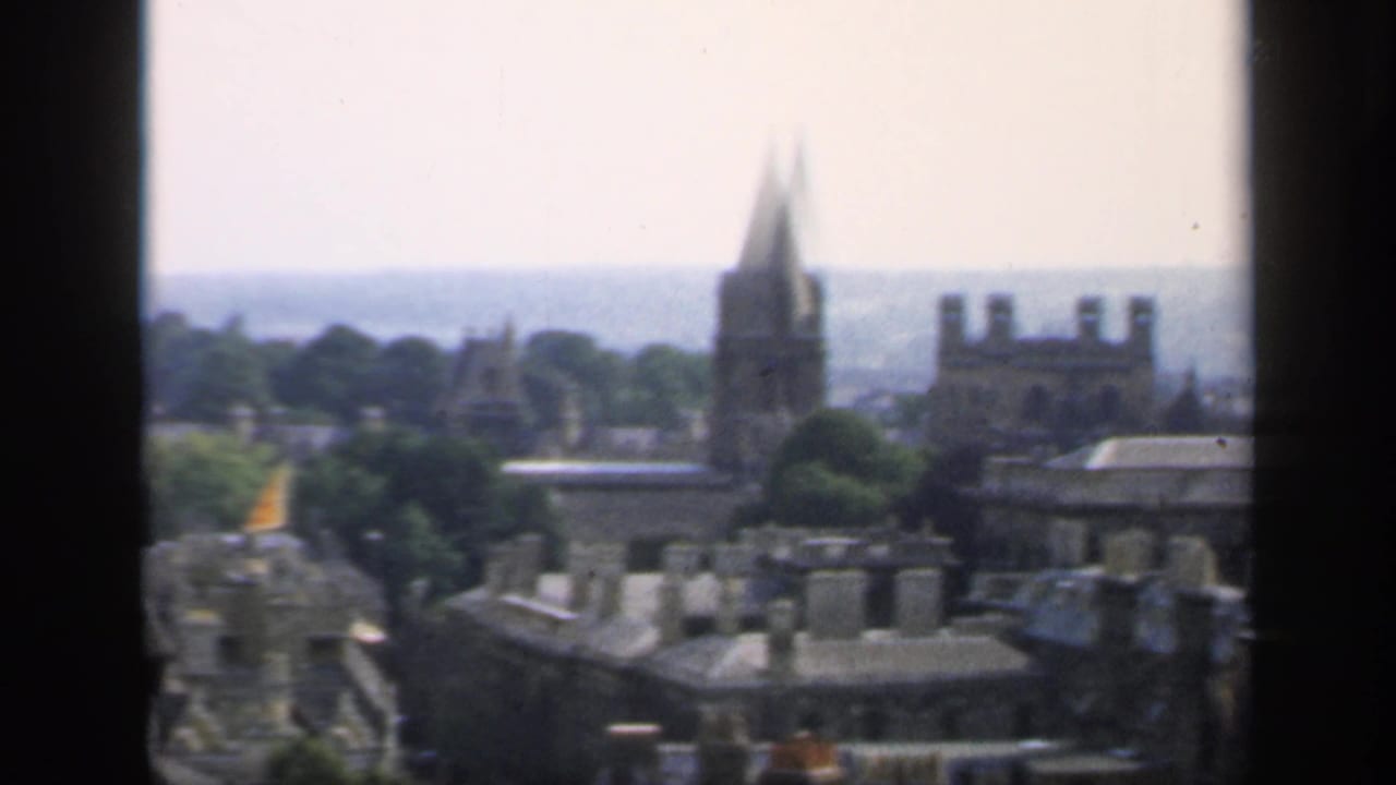 1949 8mm view over an English city, with stone rooftops, chimney stacks, and a tall spire framed by dark window edges.