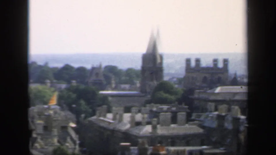 1949 8mm view over an English city, with stone rooftops, chimney stacks, and a tall spire framed by dark window edges.