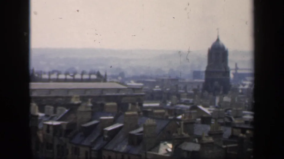 1949 home-movie frame looking across English rooftops toward a domed tower, with chimneys and distant structures softened by haze.