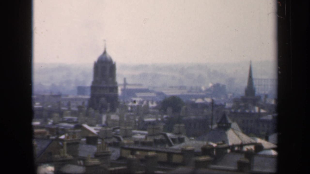 1949 England home-movie frame showing rooftops, chimneys, a domed tower, and distant church spires through window mullions.