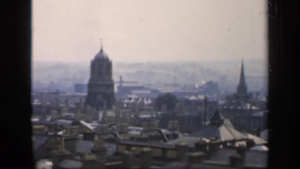 1949 England home-movie frame showing rooftops, chimneys, a domed tower, and distant church spires through window mullions.