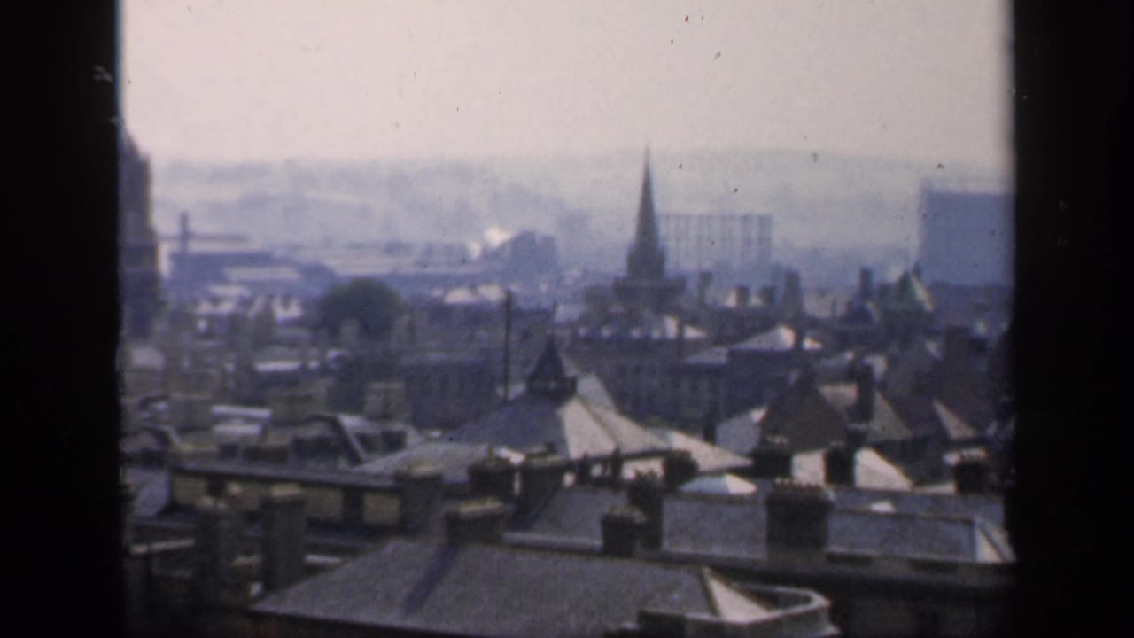 Hazy 1949 England cityscape seen from an upper window, with clustered roofs, spires, and distant buildings.