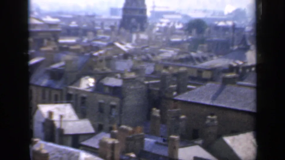 1949 England rooftop scene with slate roofs, chimney pots, narrow streets, and a distant domed tower through haze.
