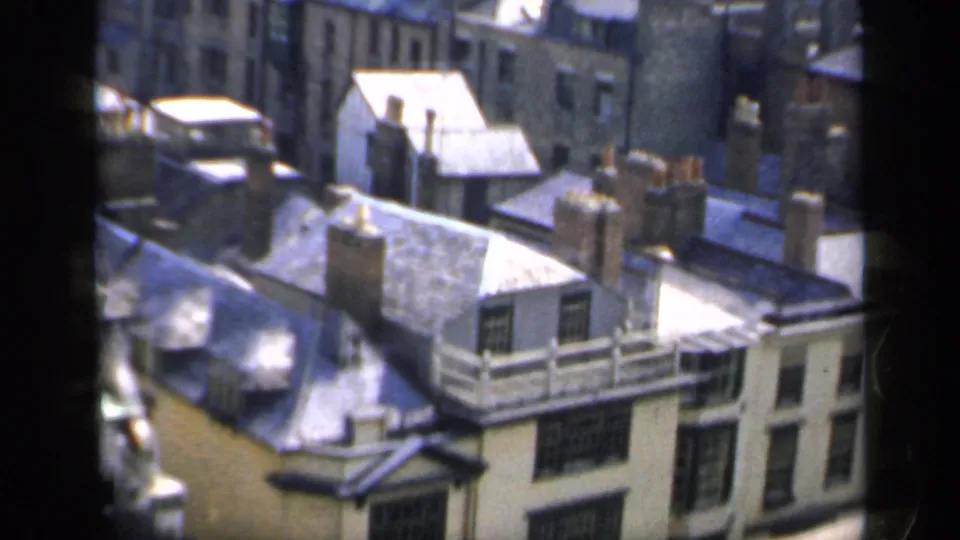 1949 England frame looking down over rooftops, chimneys, and older building facades from behind window mullions.