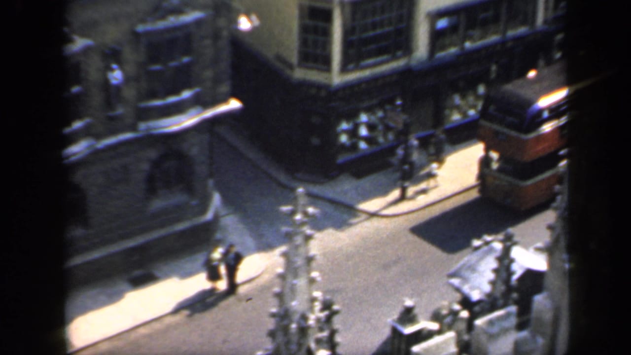 1949 England 8mm frame showing a red double-decker bus on a narrow urban street beside stone buildings and pedestrians.