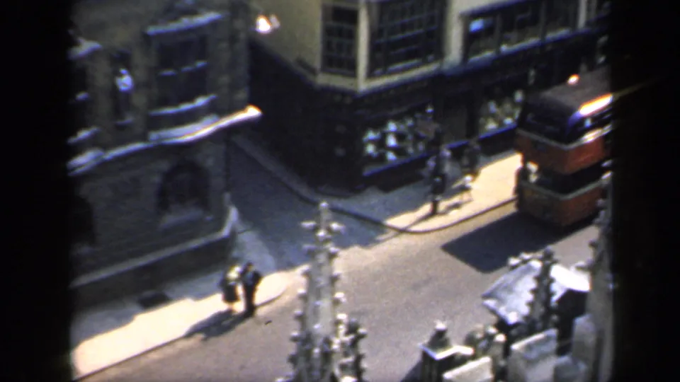 1949 England 8mm frame showing a red double-decker bus on a narrow urban street beside stone buildings and pedestrians.