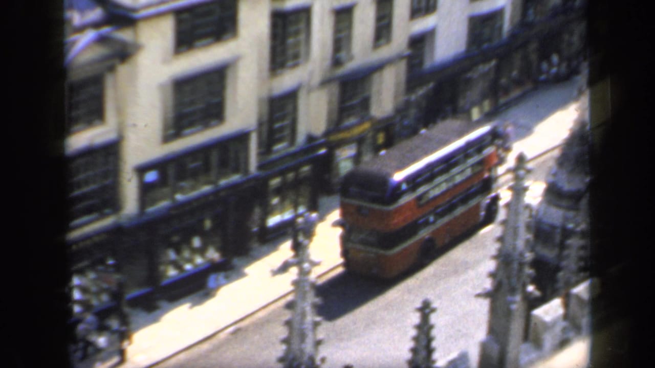 1949 England street view with a red double-decker bus, pedestrians, and commercial-looking ground-floor windows.