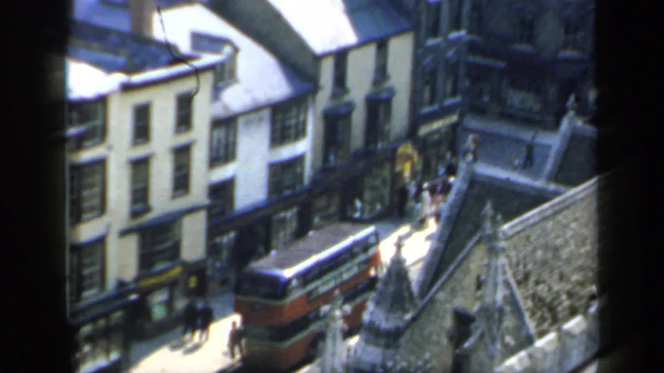 Elevated 1949 England home-movie view of a red bus below historic-looking buildings and pedestrians on the pavement.