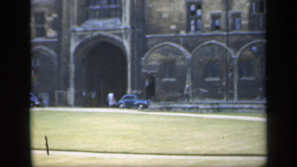 1949 England 8mm frame showing a stone building with arched entrances, a grassy foreground, and a small blue car.