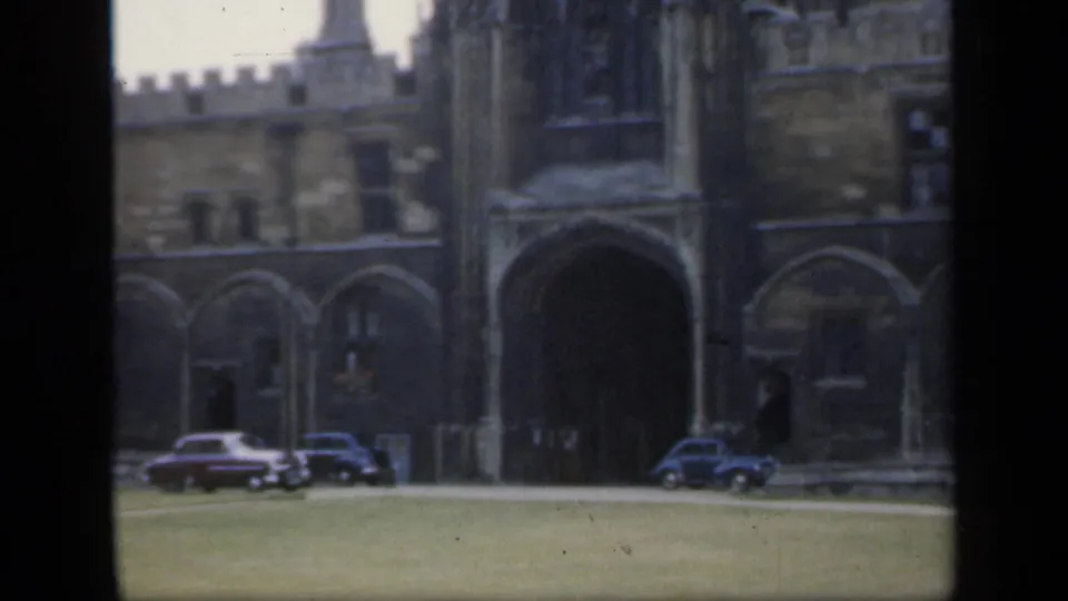 1949 England home-movie view of a historic stone courtyard facade, green lawn, arched entrance, and several parked period cars.