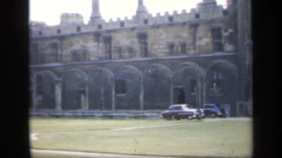 1949 England frame showing a historic stone arcade and lawn, with vintage cars parked near the building under overcast light.