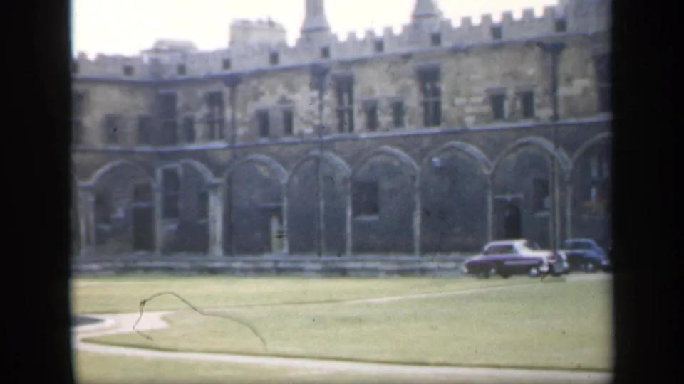 1949 England home-movie frame showing a Gothic-style courtyard facade, grass lawn, curved path, and parked mid-century cars.