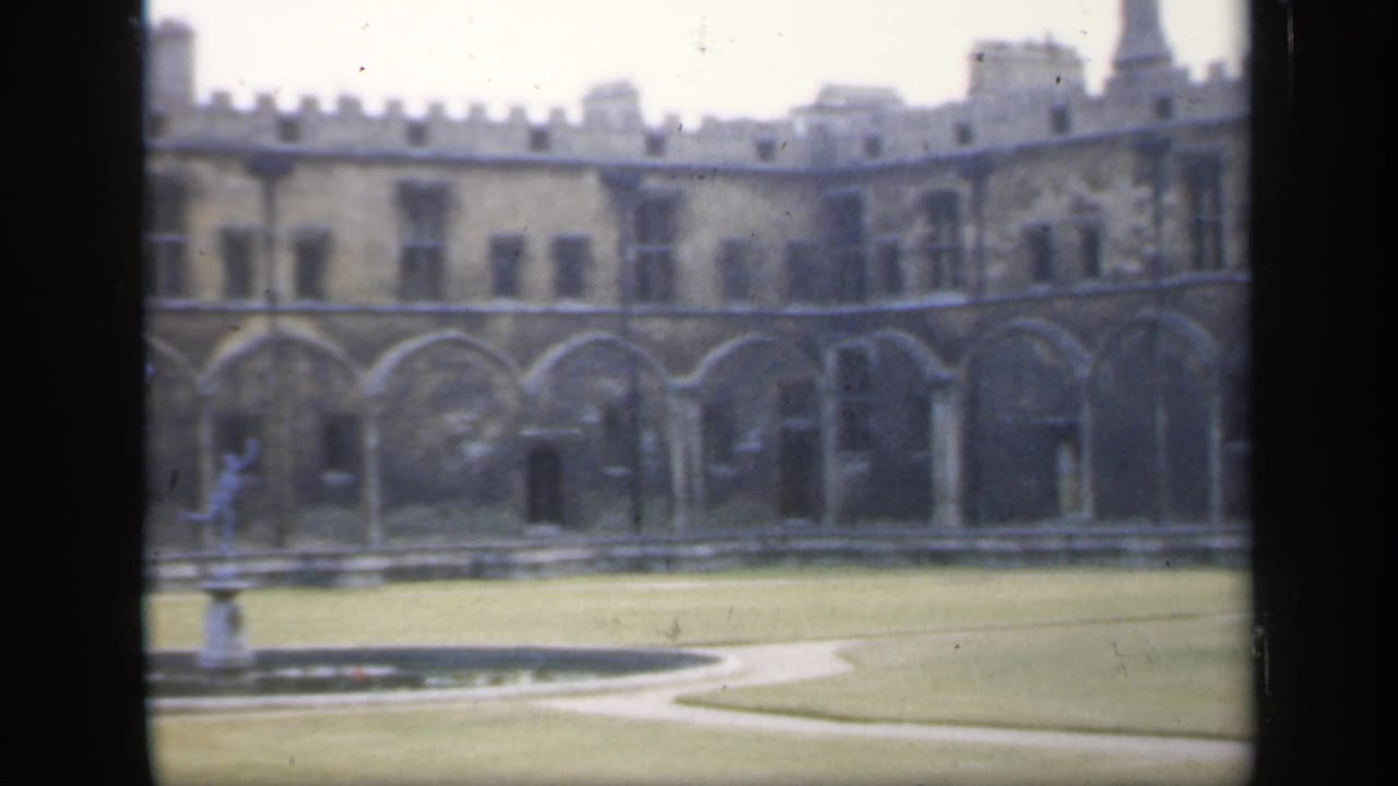Courtyard lawn with fountain statue before a long arched stone building.