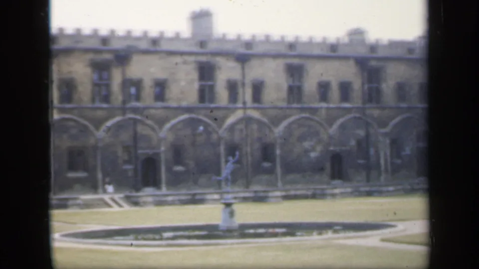1949 England home-movie view of a courtyard fountain, lawn, and repeated stone arches beneath battlements.
