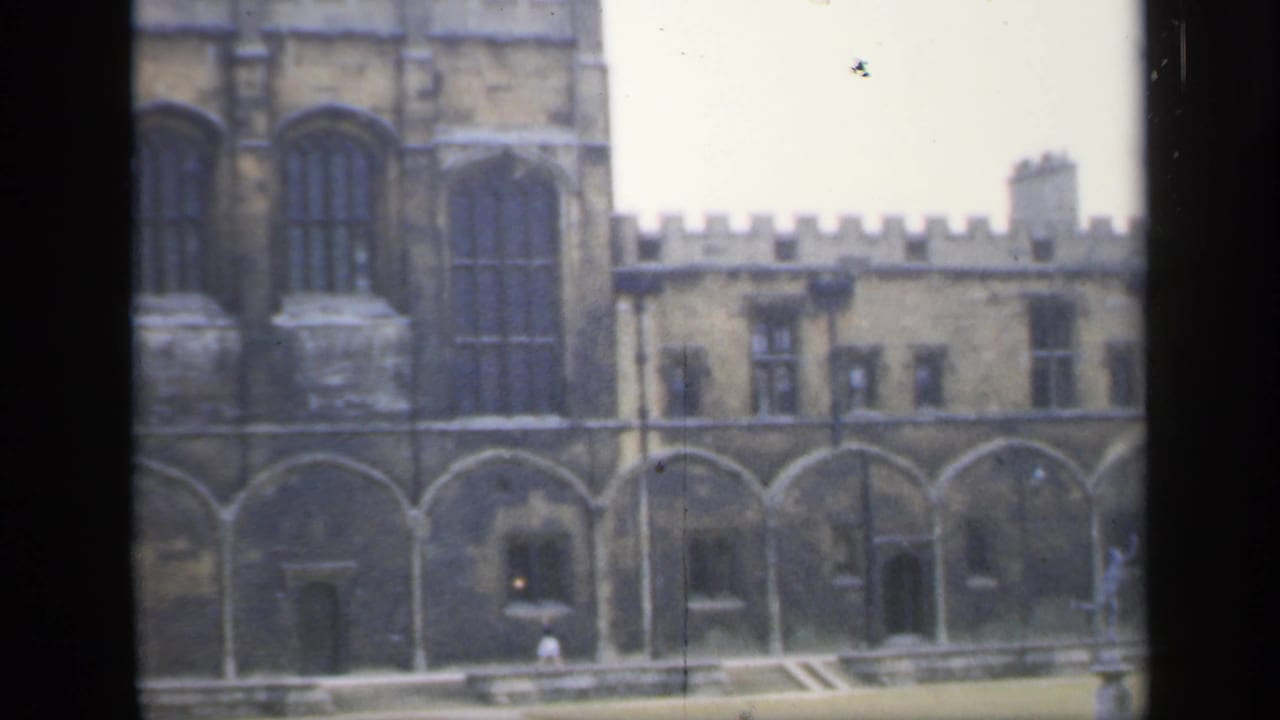 1949 England home-movie frame showing a castle-like stone building with Gothic-style windows, crenellations and courtyard details viewed through dark framing.