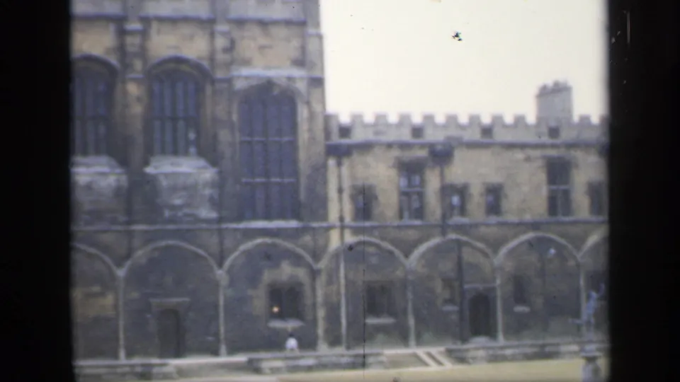 1949 England home-movie frame showing a castle-like stone building with Gothic-style windows, crenellations and courtyard details viewed through dark framing.