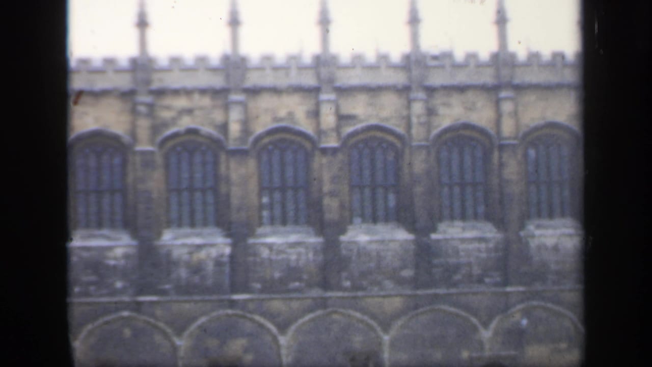 1949 England home-movie frame showing a historic stone building facade with tall arched windows and crenellated battlements.