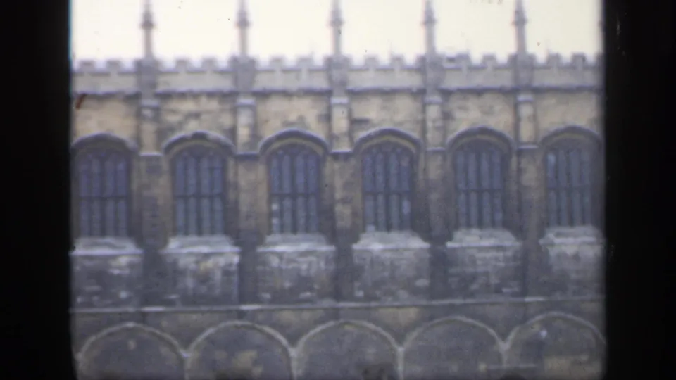 1949 England home-movie frame showing a historic stone building facade with tall arched windows and crenellated battlements.