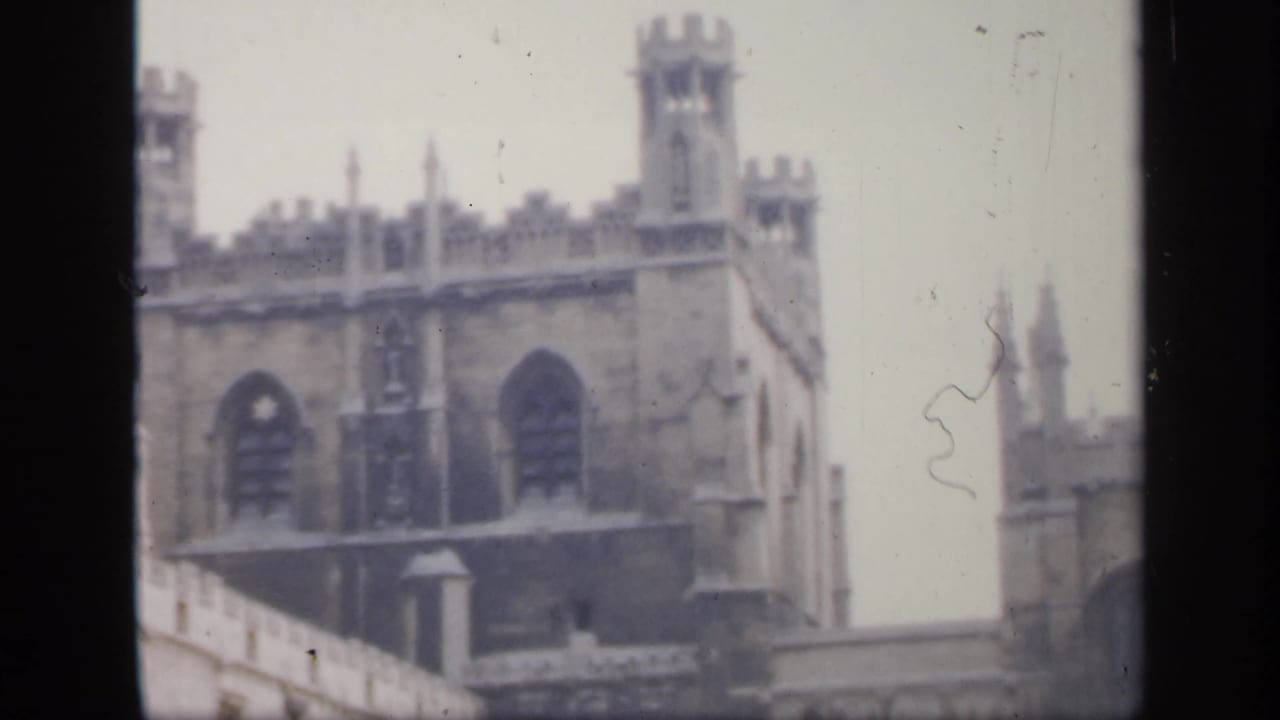 1949 England home-movie frame showing Gothic stone towers, tall arched windows, and part of a courtyard wall in soft focus.