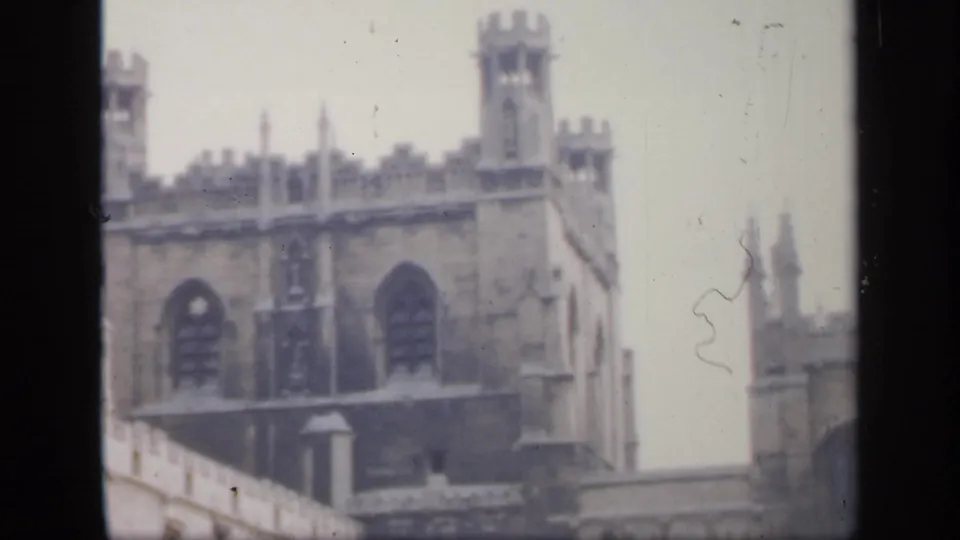 1949 England home-movie frame showing Gothic stone towers, tall arched windows, and part of a courtyard wall in soft focus.