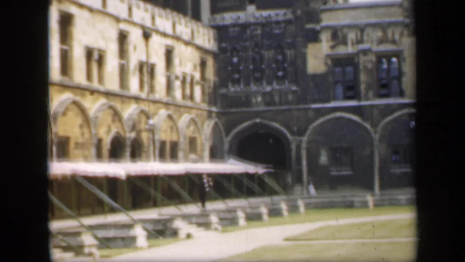 1949 England frame showing a Gothic courtyard, arched stone walkways, a lawn, and small distant visitors for scale.